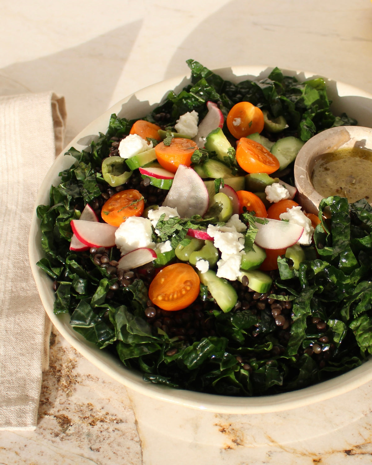 A close-up shot of a fresh kale salad in a white bowl, featuring dark green kale leaves, sliced orange cherry tomatoes, white radishes, crumbled white cheese, and cucumber, with a small wooden side dish of dressing on a light stone surface.